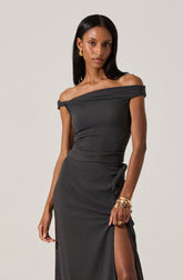 A woman is wearing an elegant off-the-shoulder black dress with a textured fabric. The dress has a draped neckline that sits off her shoulders and fits closely at the waist, flowing down to the floor with a high slit on one side. She accessorizes with gold earrings and multiple gold bracelets on her left wrist.